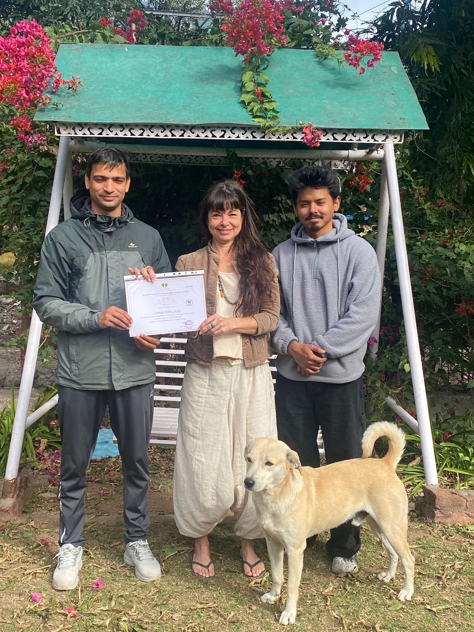 L'instructrice de yoga reçoit son diplôme en Inde, accompagnée de deux autres personnes et d'un chien dans un jardin verdoyant.